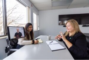 Two co-workers sit at a meeting table while a third person joins by video call on a screen, and one person communicates using sign language.